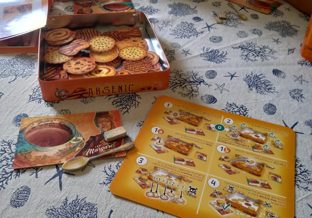 Scatola del gioco "Tea Scones and Arsenic aperta con i biscotti, e dotazione giocatore con plancia, cucchiaino e zollette