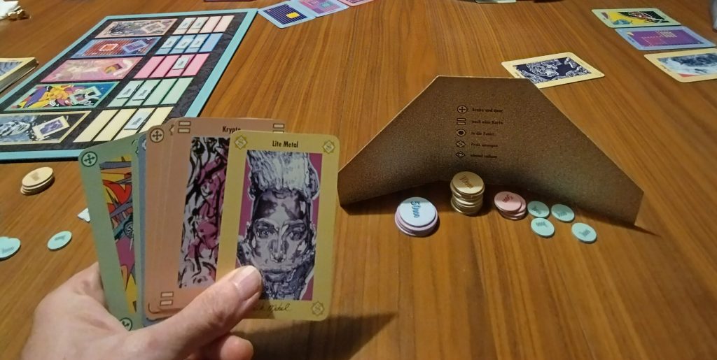 Mano di carte di Modern Art tra cui il banditore può scegliere e gettoni accumulati durante la partita dietro lo schermo giocatore